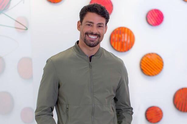 Cauã Reymond de camisa manga longa verde musgo, com as mãos nos bolsos, sorrindo, durante gravação nos estúdios do Canal Brasil