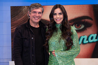 Chef Oliver Anquier de camisa preta, sorrindo, usando óculos de grau, abraçado com Daniela Albuquerquer, de vestido verde, cabelo solto, sorrindo, durante gravação do Sensacional, da RedeTV!