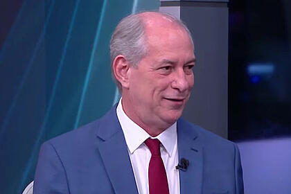 Ciro Gomes de palitó azul, gravata vermelha e camisa social branca, sentado durante entrevista na CNN Brasil