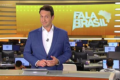 Eduardo ribeiro, de terno sem gravata, atrás da bancada nos estúdios do fala brasil, com a redação de jornalismo ao fundo e o telão com o logo do telejornal