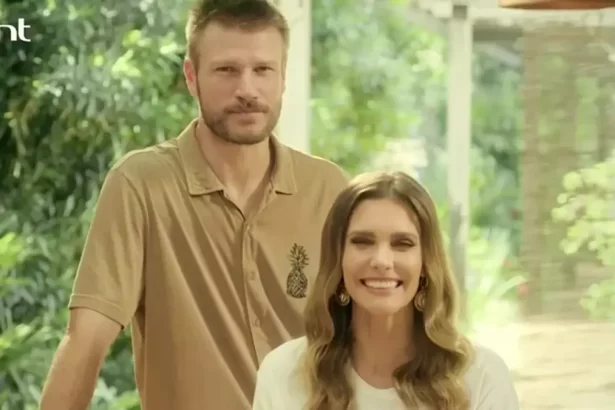 Rodrigo Hilbert com semblante sério, camisa polo bege/marrom, atrás de Fernanda; Fernanda Lima de camisa branca, sorrindo, sentada, durante gravação do Bem Juntinhos, do GNT