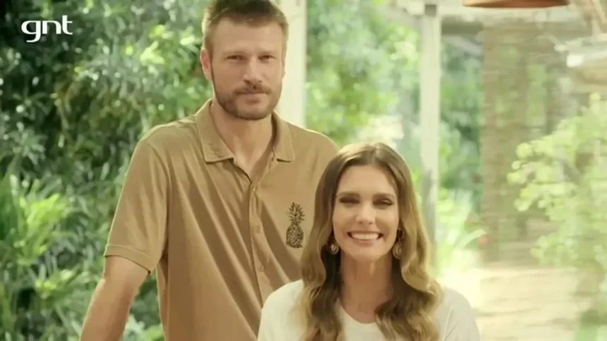 Rodrigo Hilbert com semblante sério, camisa polo bege/marrom, atrás de Fernanda; Fernanda Lima de camisa branca, sorrindo, sentada, durante gravação do Bem Juntinhos, do GNT