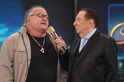 Guilherme arantes e raul gil no palco do programa raul gil. Raul segura o microfone dourado próximo a boca de guilherme