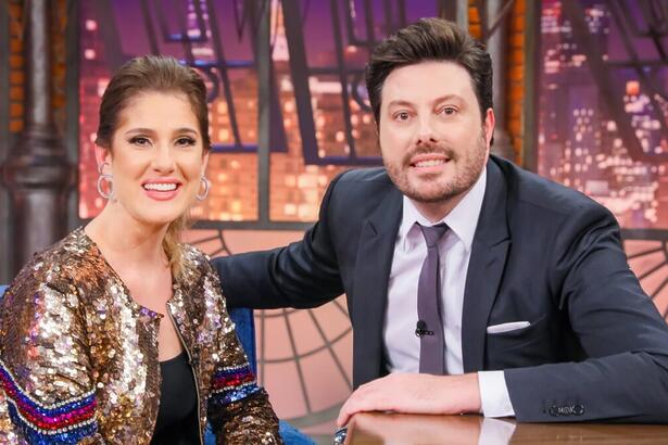 Karina Dohme de cardigã de paetê dourado e listras vermelha e roxa, camisa regata preta, sorrindo, sentada num sofá azul ao lado de Danilo Gentili durante gravação do The Noite, do SBT