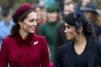 Kate Middleton de boina e sobretudo magenta ao lado de Meghan Makle de boina e sobretudo preto