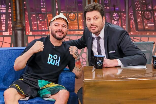 Rony Jason de short preto com estampa em verde e amarelo, camisa preta estampada na cor azul, fazendo um soco com as duas mãos e a máscara na cabeça; Danilo Gentili de palitó, gravata e calça preta e camisa social branca; ambos sentados durante gravação do The Noite do SBT