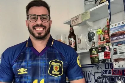 Felippe Facincani com a camsa de um clube, em frente a uma preteleeira com troféus e livros