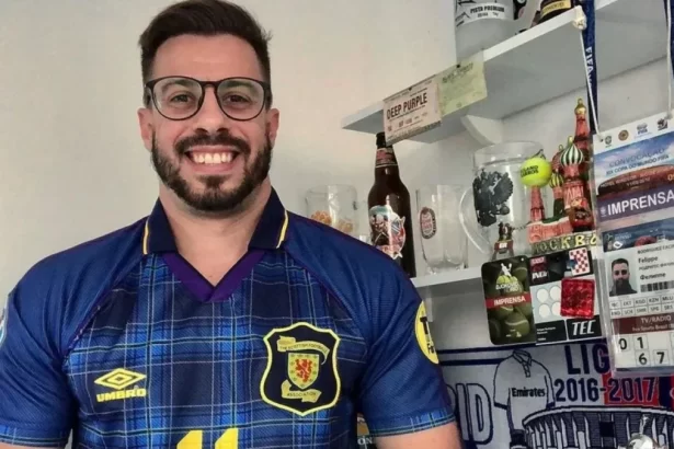 Felippe Facincani com a camsa de um clube, em frente a uma preteleeira com troféus e livros