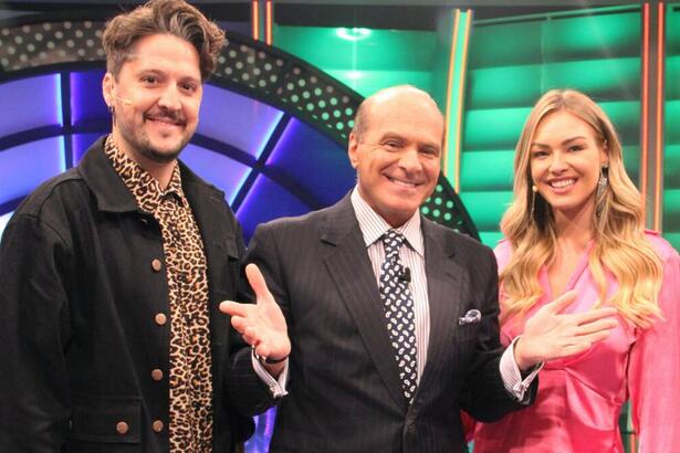 André Vasco de jaqueta preta e camisa estampada sorrindo; Marcelo de Carvalho sorrindo, de braços abertos, de palitó preto e gravata cinza com branco; Barbara Heck sorrindo, de vestido decotado rosa durante gravação de O Céu é o Limite, da RedeTV!