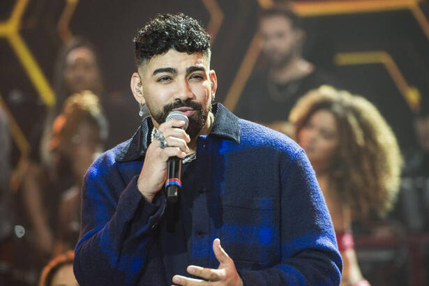 Dilsinho de camisa social azul e preta, segurando o microfone preto com a mão direita durante performance no Faustão da Band