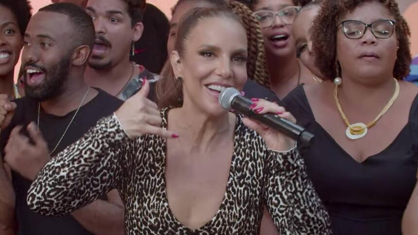 Ivete Sangalo de macacão animal-print fazendo um telefone com a mão direita segurando um microfone com a mão esquerda durante gravação do Macaco Sessions