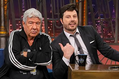 Joel Santana de camisa e calça preta, semblante sério, braços cruzados, ao lado de Danilo Gentili durante gravação do The Noite, do SBT