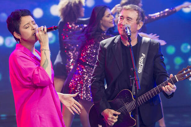 Kell Smith de rosa e Amado Batista de blazer e camisa pretos segurando uma guitarra, cantando durante gravação do Faustão na Band