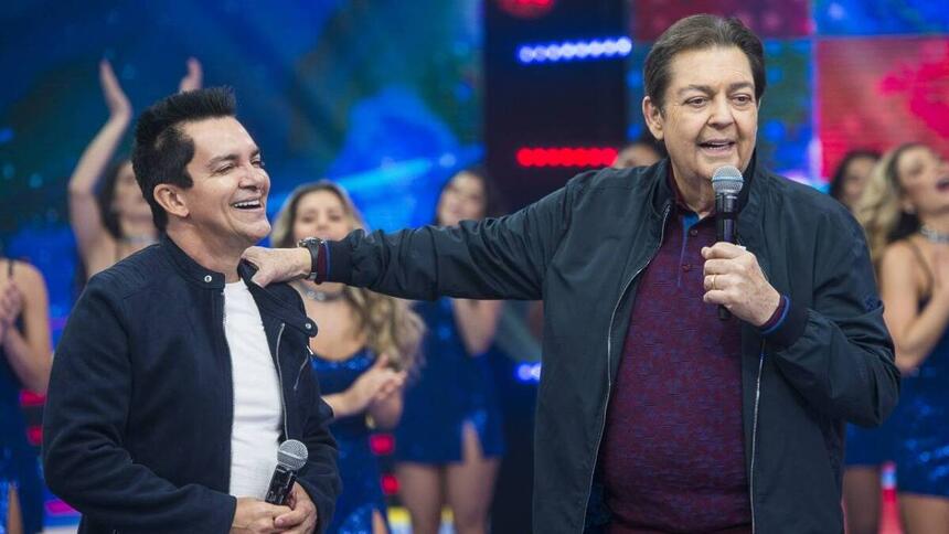 Regis Danese sorrindo, de camisa branca e preta, ao lado de Faustão durante gravação de programa