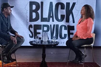 Seu Jorge e Tábata Poline sentados durante entrevista para o Fantástico, da TV Globo