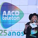 Adriane Galisteu de chapéu de Cowgirl preto, segurando fichas e microfone durante apresentação do Teleton