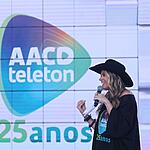 Adriane Galisteu de chapéu de Cowgirl preto, segurando fichas e microfone durante apresentação do Teleton