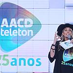 Adriane Galisteu de chapéu de Cowgirl preto, segurando fichas e microfone durante apresentação do Teleton