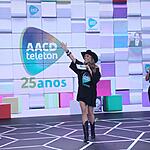 Adriane Galisteu de chapéu de Cowgirl preto, segurando fichas e microfone durante apresentação do Teleton