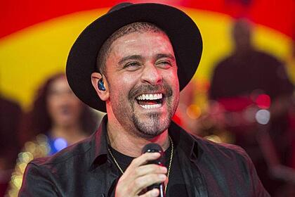 Diogo Nogueira sorrindo, de chapéu e camisa preta, segurando um microfone com a mão direita durante performance no Na Pista do Sucesso, do Faustão na Band