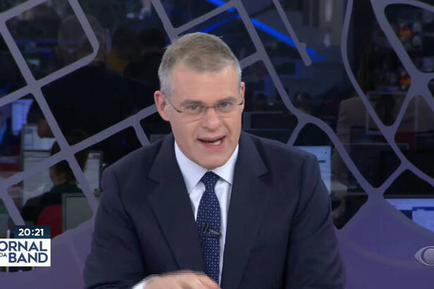 Eduardo Oinegue de paletó e gravata pretos, sentado durante apresentação do Jornal da Band