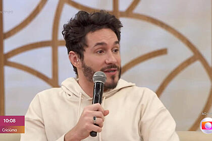 Eliezer com um moletom branco, olhando para o seu lado esquerdo no sofá do Encontro. Ele segura um microfone na mão