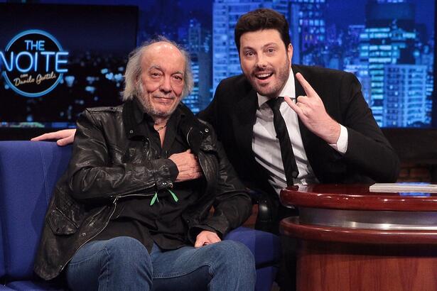 Erasmo Carlos sorrindo, de blazer de couro preto, calça jeans, sentado num sofá azul ao lado de Danilo Gentili, durante gravação do The Noite, do SBT