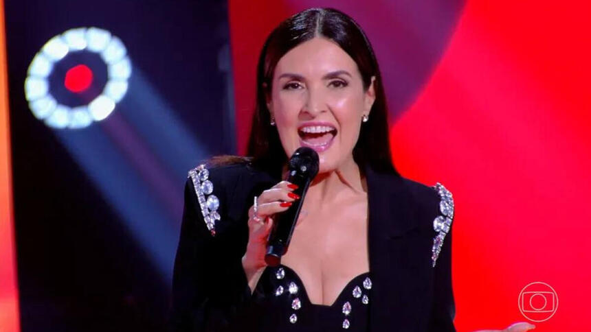 Fátima Bernardes no estúdio do The Voice Brasil. Ela usa uma roupa preta e um microfone de mão