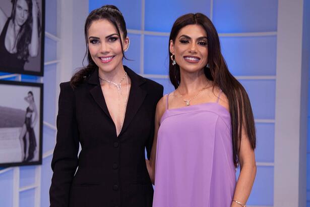 Daniela Abuquerque de vestido decotado preto, cabelo preso, sorrindo, ao lado de Flávia Noronha com vestido roxo, cabelo solto, sorrindo durante gravação do Sensacional, da RedeTV!
