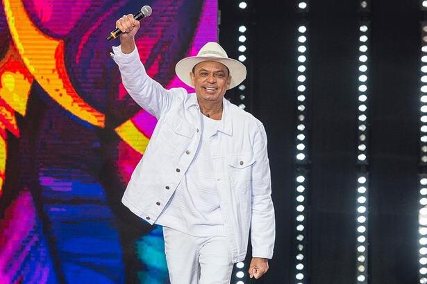 Frank Aguiar de camisa, jaqueta e calça jeans brancos, sorrindo, com o braço direito levantado durante gravação do Faustão na Band