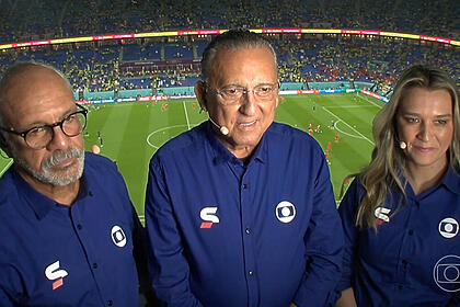 Galvão Bueno, Júnior e Ana Thaís Matos na cabine de transmissão, com o uniforme azul de esporte da Globo, com o estádio ao fundo
