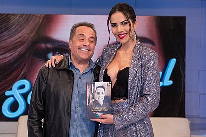 Geraldo Magela sorrindo, de jaqueta preta de couro, camisa social azul marinho ao lado de Daniela Albuquerque, que está sorrindo, segurando o livro de Geraldo, durante gravação do Sensacional, da RedeTV!