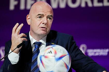 Gianni Infantino de palitó preto, gravata listrada azul e preta, com uma bola na mesa falando ao microfone durante coletiva da Copa do Mundo do Catar