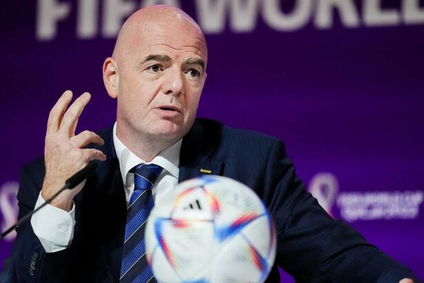 Gianni Infantino de palitó preto, gravata listrada azul e preta, com uma bola na mesa falando ao microfone durante coletiva da Copa do Mundo do Catar
