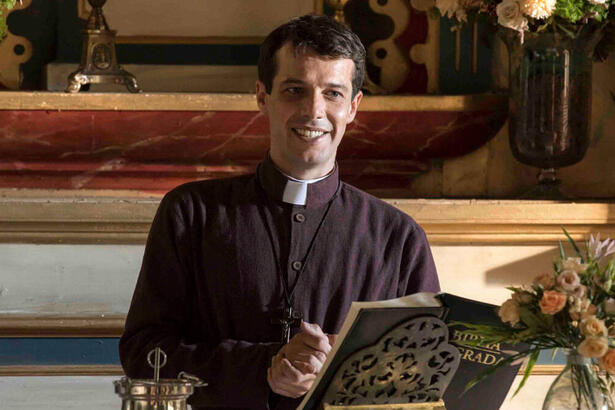 Jayme Matarazzo, como Padre Tenório, em trecho da novela Além da Ilusão. Ele veste uma batina de padre, com um sorriso no rosto, celebrando uma missa. A sua frete está uma bíblia apoiada e um arranjo de flor