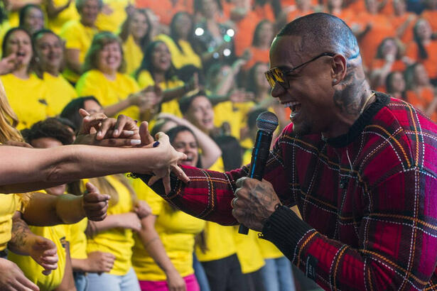 Leo Santana de camisa quadriculada em tons de preto e magenta, sorrindo, segurando a mão de uma fã na plateia do Faustão na Band