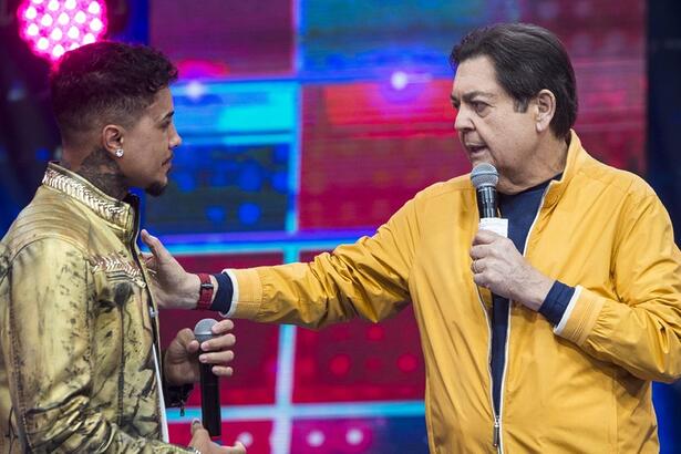 Livinho de jaqueta estampada segurando o microfone com a mão esquerda olhando pra Faustão durante gravação de programa
