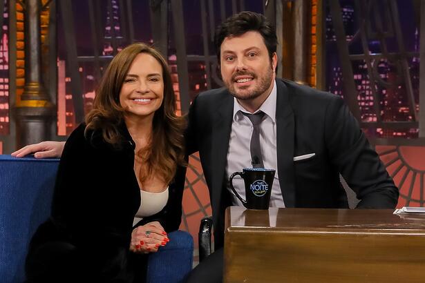 Luiza Tomé de blazer e calça preta, camisa branca por dentro, sorrindo, sentada num sofá azul ao lado de Danilo Gentili durante gravação do The Noite, do SBT