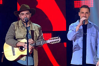 Dupla Neto & Felipe cantando no palco do The Voice Brasil. O primeiro toca um violão e tem um chapéu na cabeça. O segundo segura um microfone de mão