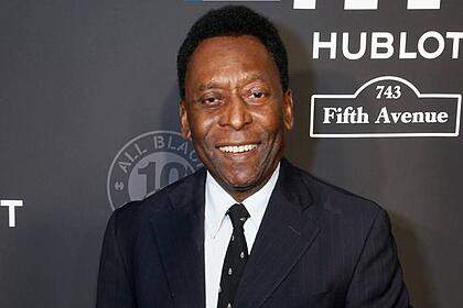 Pelé com um terno e gravata, sorrindo, em foto posada em frente e um banner preto de um evento