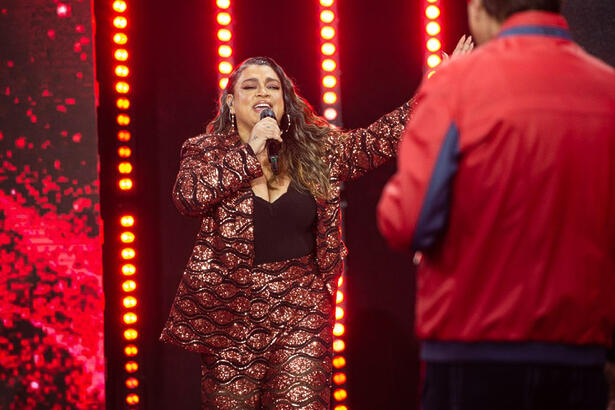 Preta Gil de blazer a calça vermelha com lantejolas, camisa preta, com os braços abertos segurando o microfone com a mão direita durante gravação do Faustão na Band