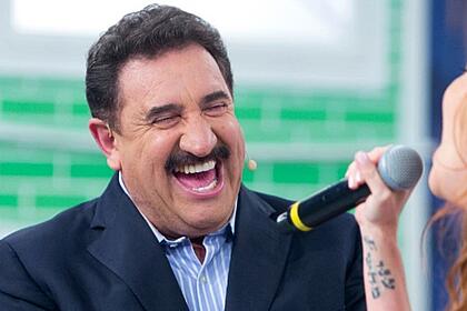 Ratinho de blazer azul escuro e camisa social azul clara, sorrindo na gravação do seu programa