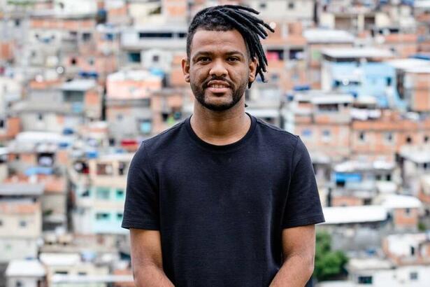 Rene Silva de camisa básica preta, sorrindo, de cabelo trançado preso sentado durante ensaio fotográfico no Morro