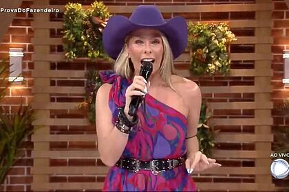 Adriane com o microfone na mão, com uma roupa estampada e um chapéu de cor lilás.