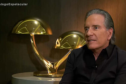 Roberto Justus com uma blusa em tom mais escuro, em trecho da entrevista para o Domingo Espetacular, em sua residência, com um enfeite dourado ao fundo