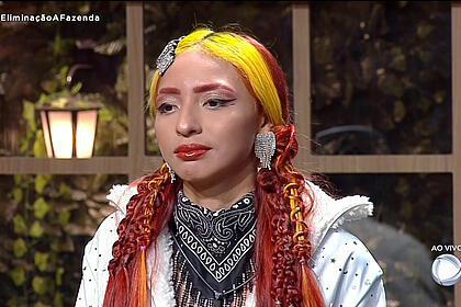 Ruivinha de Marte com camisa branca, cachecol cinza, cabelo partido ao meio, de batom vermelho, sentada na sede de eliminação de A Fazenda, da Record TV