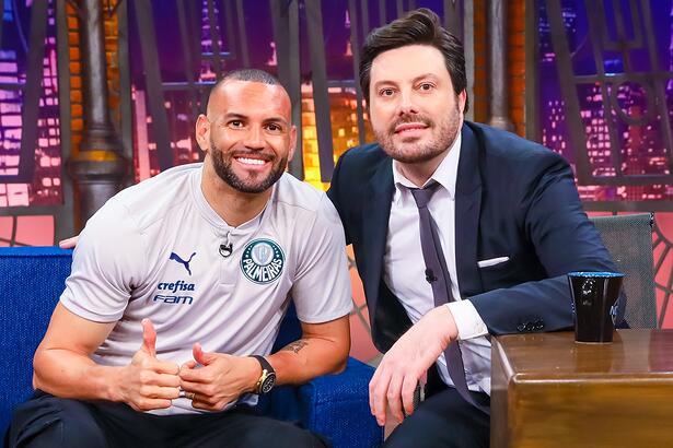 Weverton Pereira sorrindo, de camisa cinza, fazendo legal com as duas mãos, sentado ao lado de Danilo Gentili durante gravação do The Noite, do SBT
