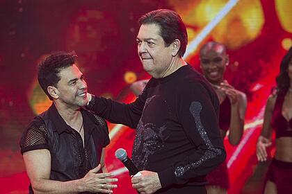 Zezé di Camargo usando camisa social preta, sorrindo, estendendo a mão esquerda pra Faustão; Faustão de camisa manga longa preta, segurando o micrfone com a mão esquerda e com a direta no pescoço de Zezé, durante gravação do Faustão na Band