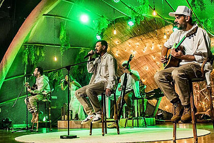 Integrantes da Planta e Raiz sentados, de camisa branca e calça marrom durante gravação do 'Acustico Planta e Raiz'