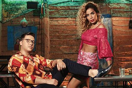 Wesley Safadão de camisa social estampada nas cores laranja, vermelho e branco, sentado, de óculos de grau, calça preta, com o pé esticado; Anitta com cabelo encaracolado, batom vermelho, saia rosa e tomara que caia em tom magenta, encostada numa mesa; ambos juntos para gravação de videoclipe